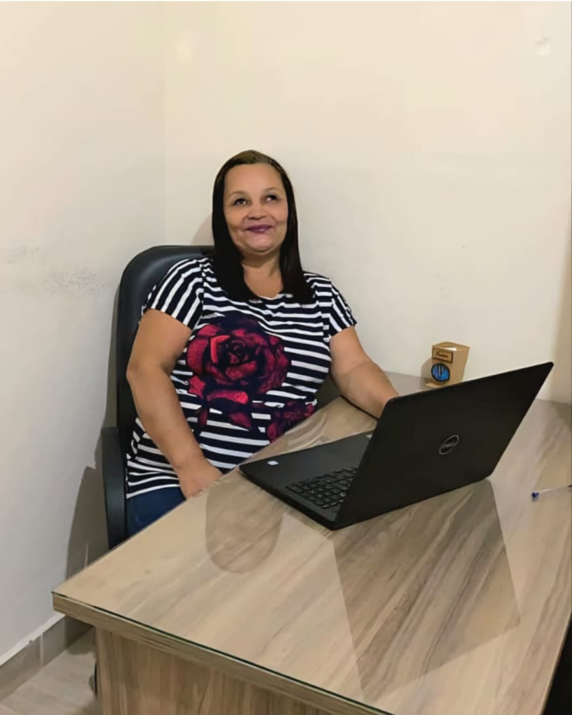 Edna Santos está sentada em uma cadeira atrás de uma mesa de madeira, sorrindo para a câmera. Ela usa uma blusa listrada em azul-marinho e branco, com uma grande estampa de rosa vermelha. Sobre a mesa há um notebook aberto, e o ambiente tem paredes claras e aparência de escritório.