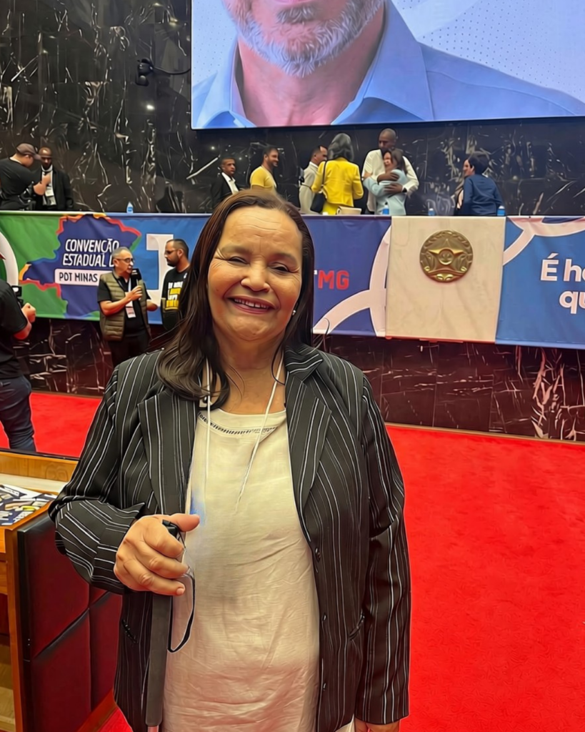 Edna Santos, sorrindo e segurando sua bengala, está em um evento político, diante de um palco com pessoas e painéis da convenção estadual do PDT de Minas Gerais. Ao fundo, há um grande telão e um tapete vermelho.
